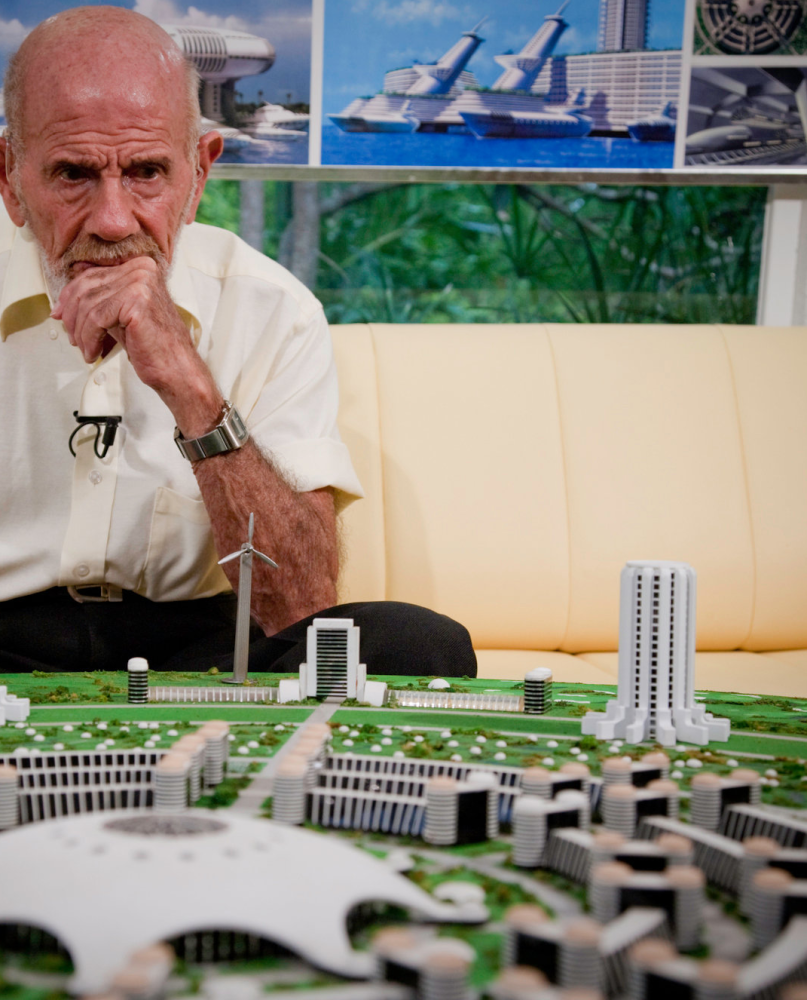 An elderly man seated behind a detailed scale model of a circular future city (ny times)