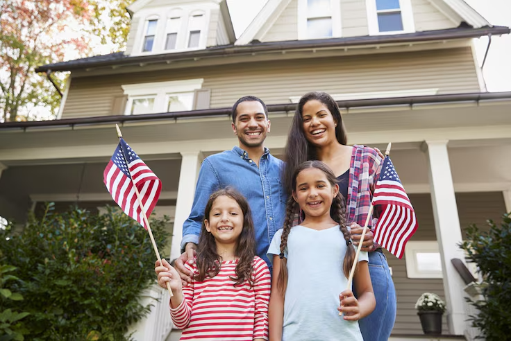 A smiling American family holding small flags in front of a large suburban house