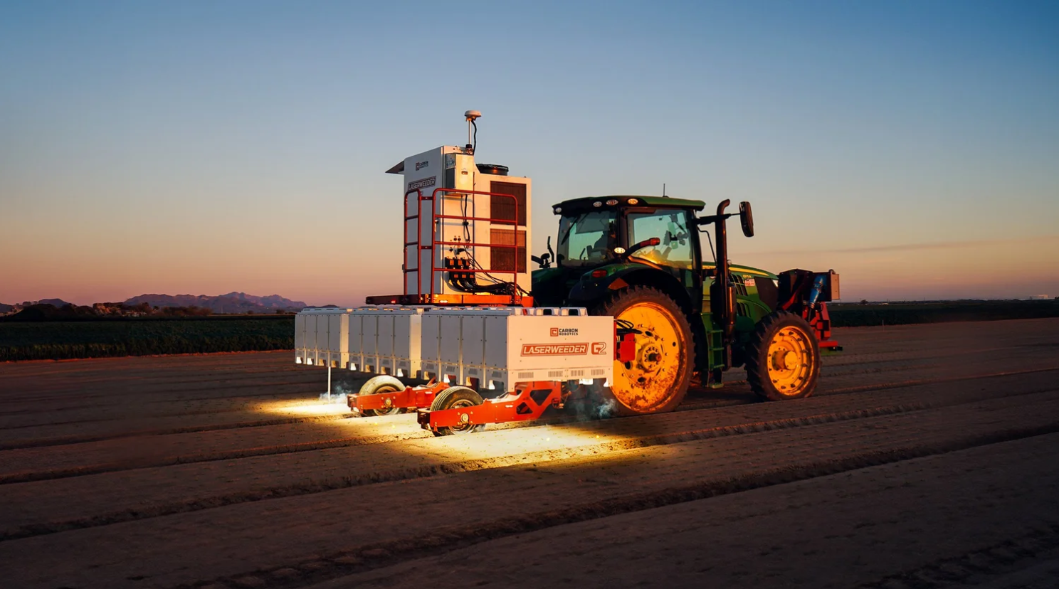 A Carbon Robotics LaserWeeder towed behind a tractor at dusk, lasers glowing over the rows